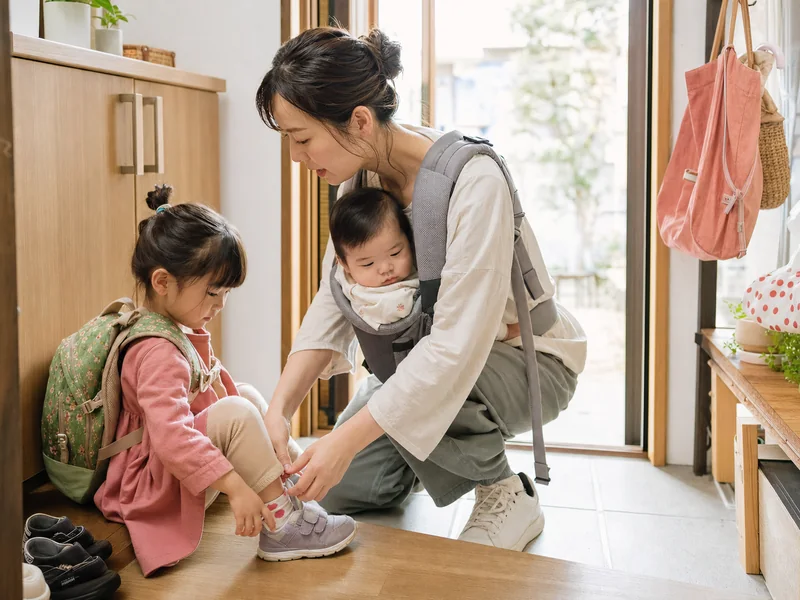 上の子の登園準備をしながら赤ちゃんを抱く日本人の母親の生活場面イメージ